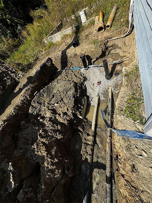Trench dug along a building's side, showing pipes and fittings in the dirt.