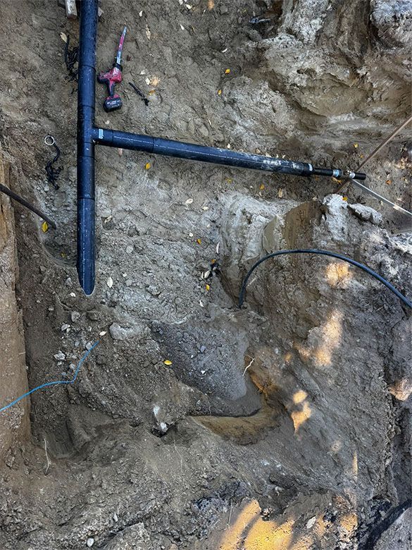 A trench in the earth holds black PVC piping; a drill and tools rest on the soil.