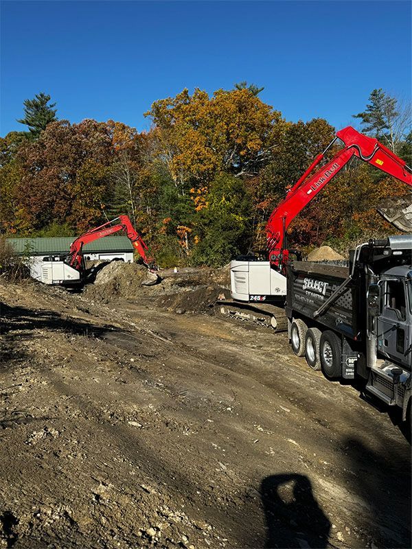 Two excavators loading a dump truck at a construction site on a sunny day with fall foliage.