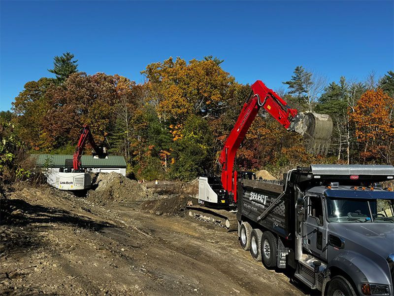 Two excavators loading dirt onto a dump truck on a construction site with fall foliage.