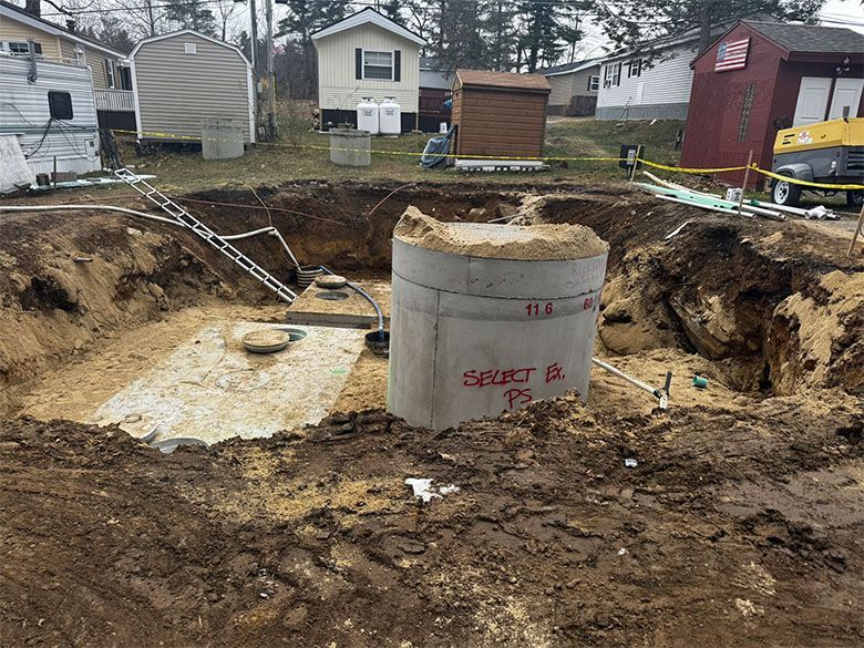 Excavation site with a concrete septic tank installed. Dirt surrounds the tank and open pit; houses in the background.
