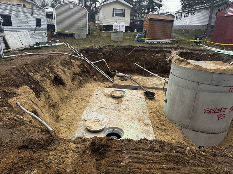 Excavation site with septic tank components, pipes, and surrounding buildings. Concrete structures are visible.