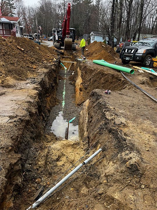 Excavation site with an excavator, a trench with piping, and a worker in a high-visibility vest.