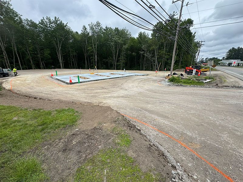 Construction site with gravel and a concrete foundation. Trees and a road visible.