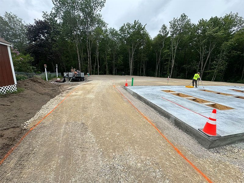 Construction site with gravel driveway leading to a concrete slab. A worker in a neon vest is present.