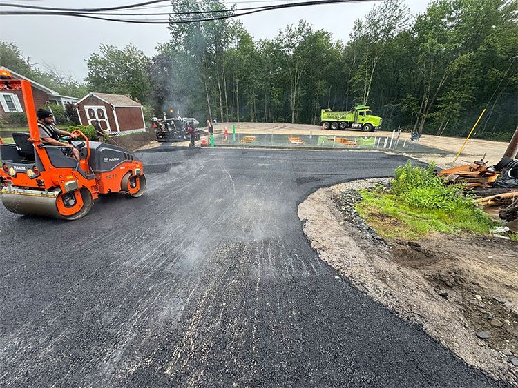 Asphalt paving with a roller machine on a street, with workers and construction vehicles nearby.