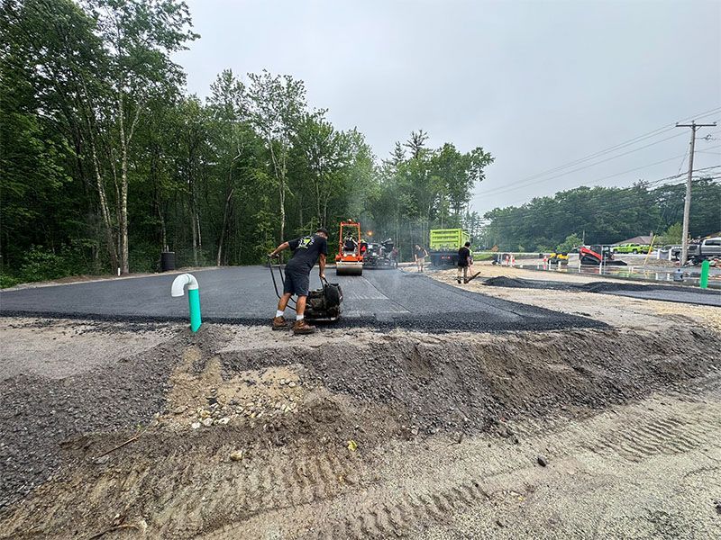Road construction site with workers laying asphalt. Grey pavement, dirt, green trees.