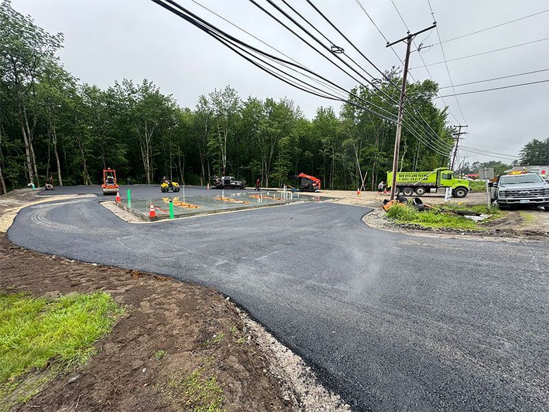 Newly paved asphalt road and entrance to a construction site. Green truck and orange construction equipment visible.