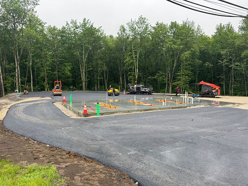 New asphalt parking lot under construction; machinery and cones present. Green trees in the background.