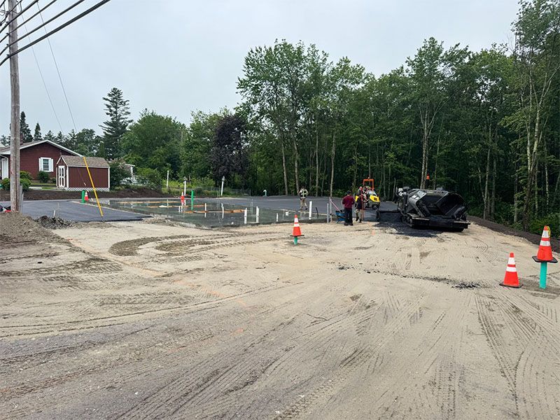 Construction site: Asphalt work. Workers near a truck, surrounded by orange cones. Trees and a small house are in the background.