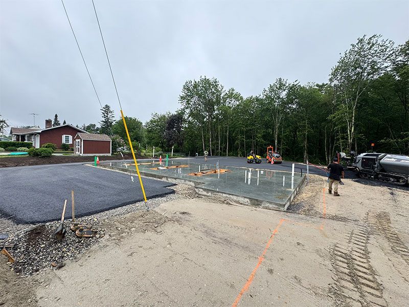 Construction site: freshly poured concrete foundation, asphalt, trees, power lines, worker.
