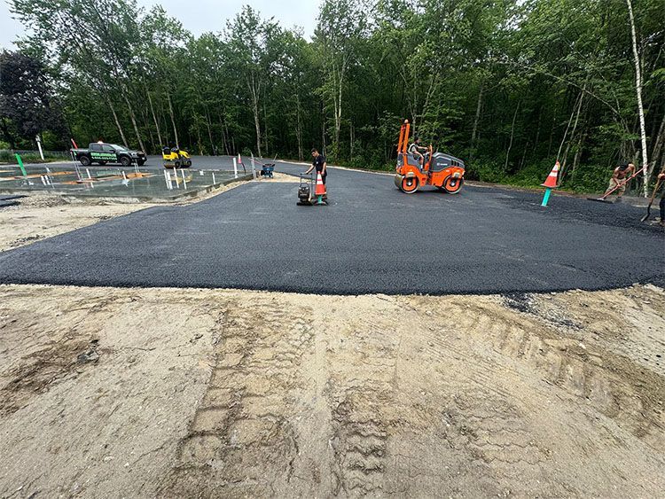 Asphalt paving in progress on a sandy lot; small roller compacting the new surface.