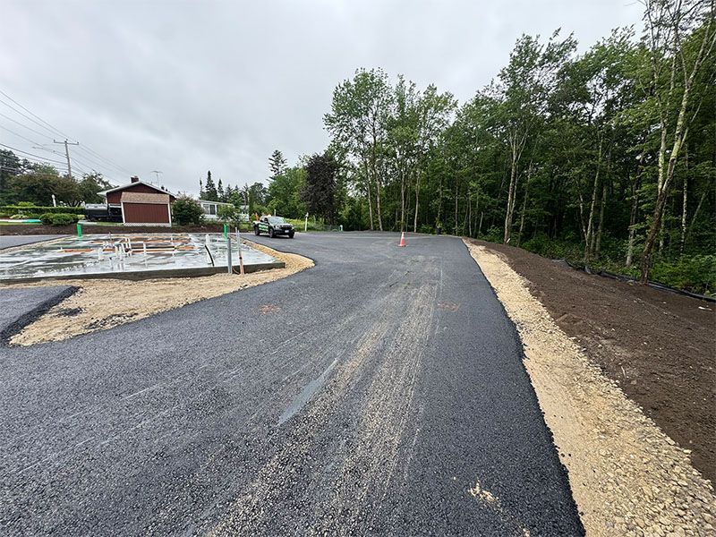 Newly paved asphalt lot with gravel edges, a few trees, and a building on a cloudy day.