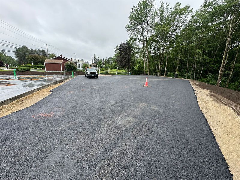 Newly paved asphalt parking lot with a vehicle, trees, and buildings in the background.