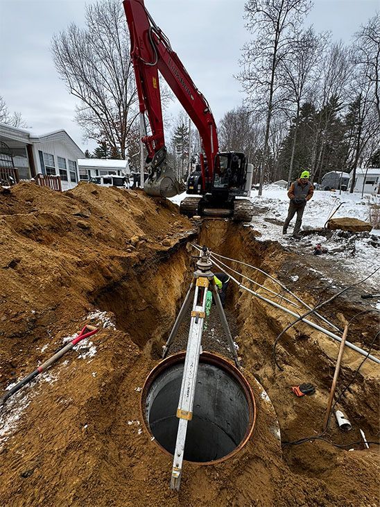 Construction site: excavator digging trench for large pipe; worker uses level. Overcast day.