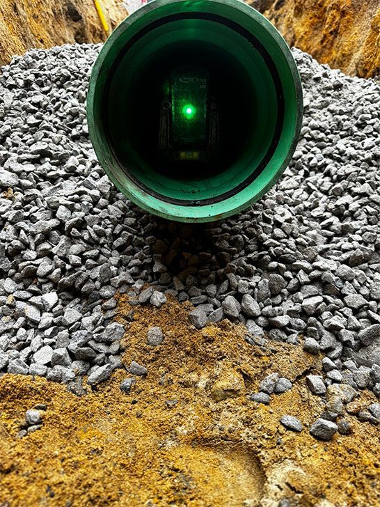 Green pipe in a trench with gravel and dirt, a green laser level inside.
