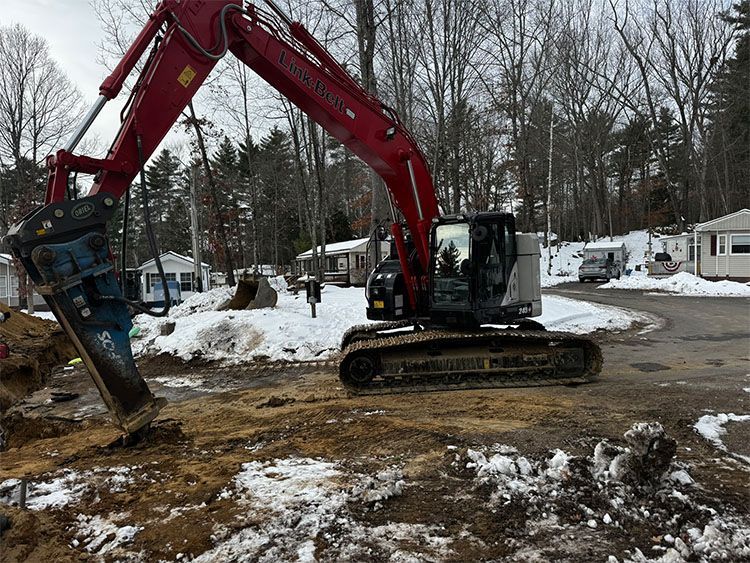 Red excavator with a jackhammer attachment on a construction site, winter setting.