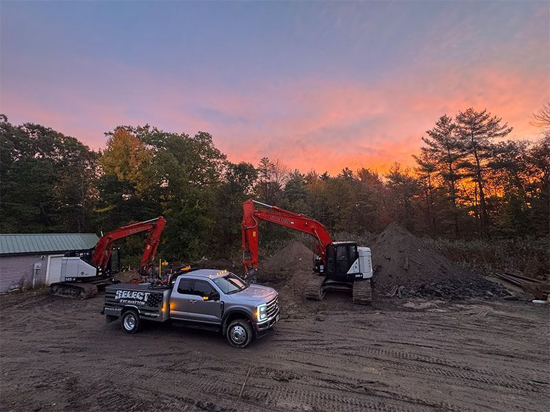A work truck with a logo, excavators and dirt pile against a colorful sunset.
