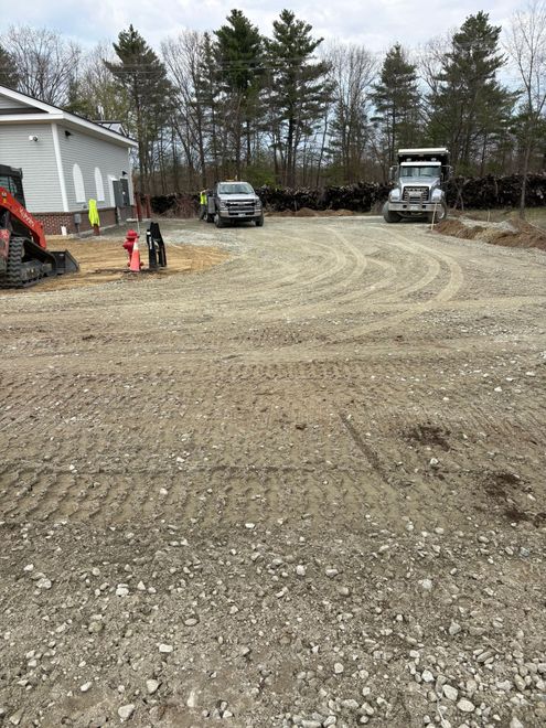 Gravel parking area under construction with trucks, machinery, and a building on the left.