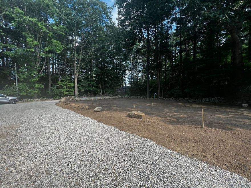 Gravel driveway leads to a cleared lot with a few rocks, surrounded by trees under a blue sky.