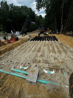Construction site with buried septic system, gravel bed, black pipes, and workers in the background.