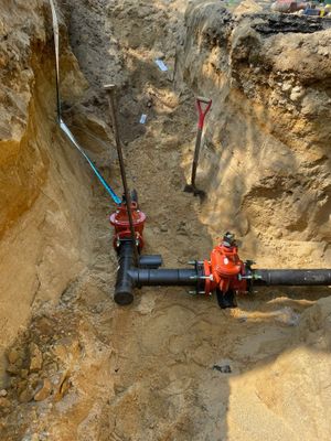 Pipes and valves in a trench, with shovels. The valves are red, the pipes black, and the soil is light brown.