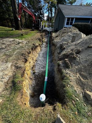 Green pipe in trench filled with gravel, by a house and excavator.