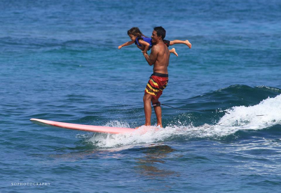 A man is holding a child on his shoulders while riding a wave on a surfboard