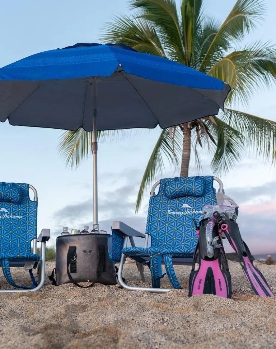 A blue umbrella is sitting on top of a sandy beach next to chairs and flippers.