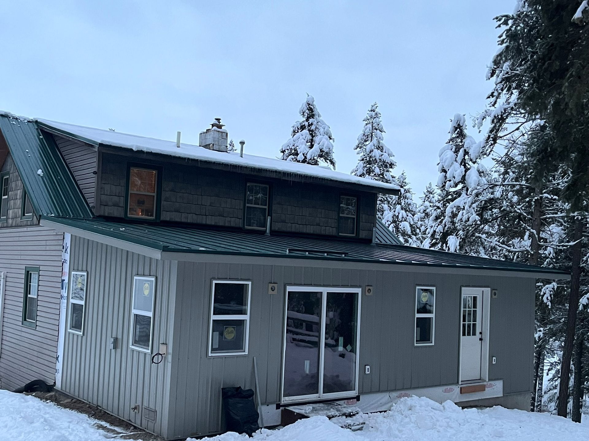 Snow-covered cabin with gray siding and a sliding glass door in a wooded winter setting