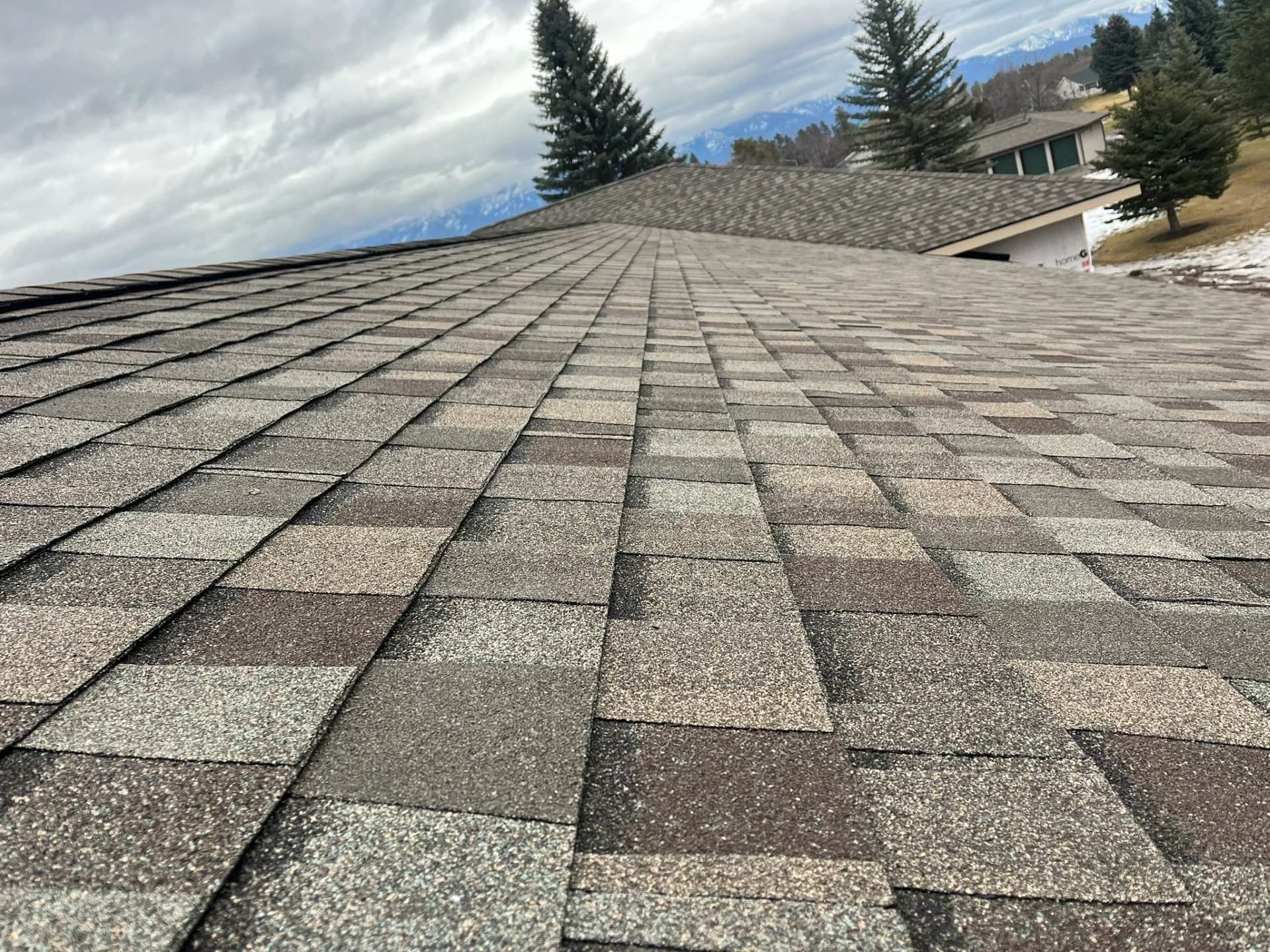Roof with gray shingles under a cloudy sky, viewed from a low angle with trees and mountains in the distance