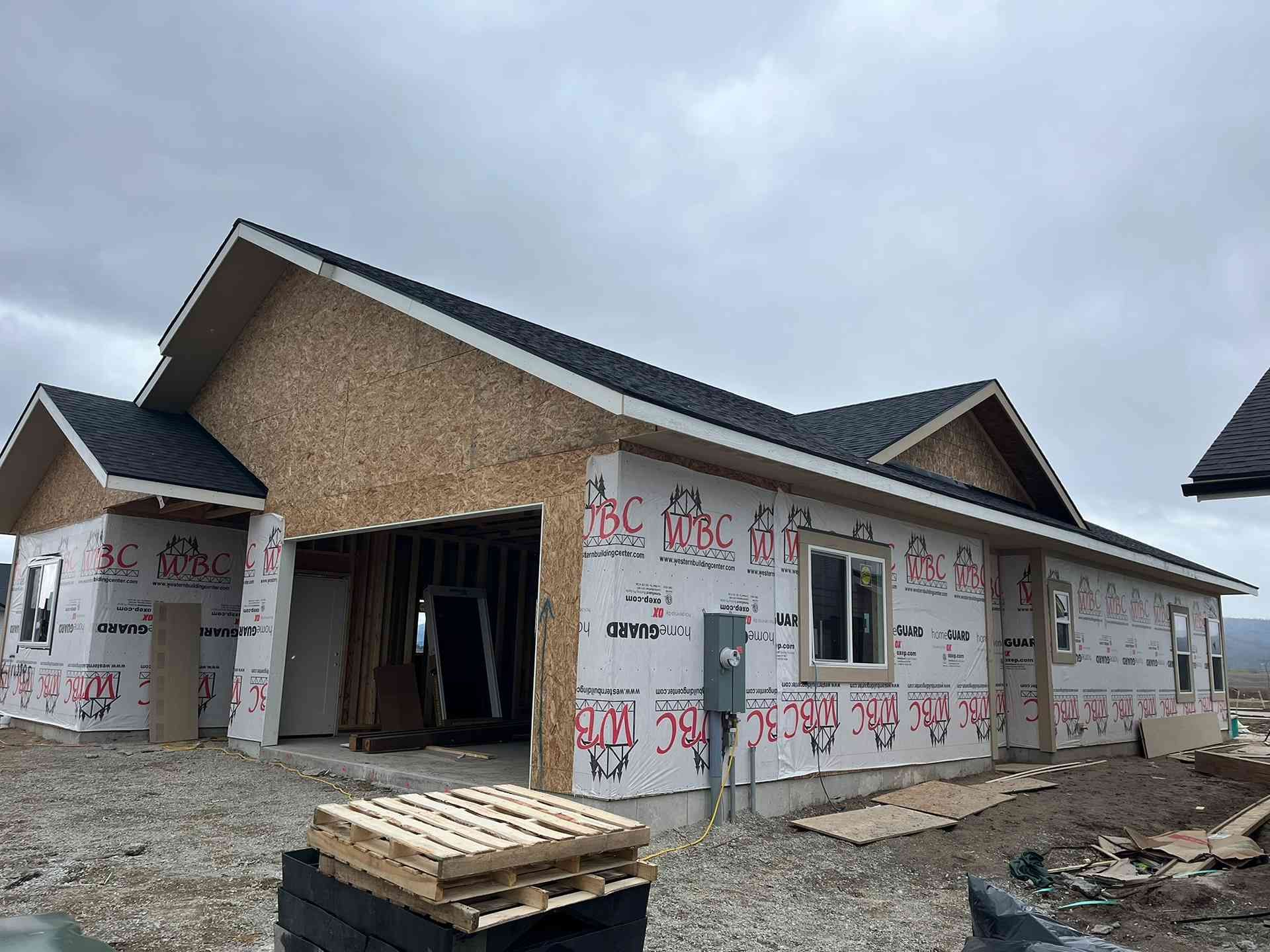 Partially built house with tan siding, white wrap, open garage, and stacked lumber under an overcast sky