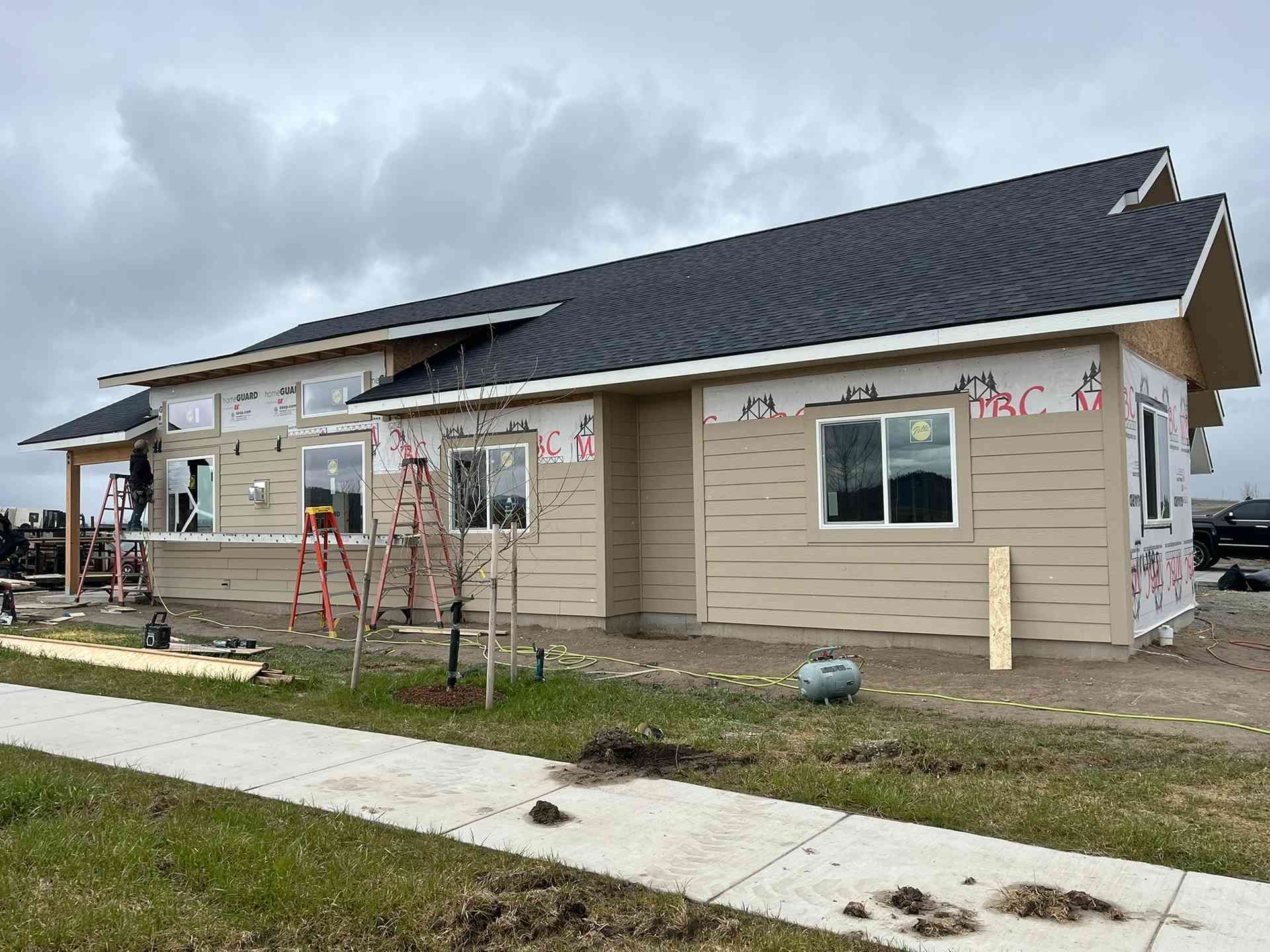 Partially built house with siding, dark roof, ladders, and construction materials under a cloudy sky