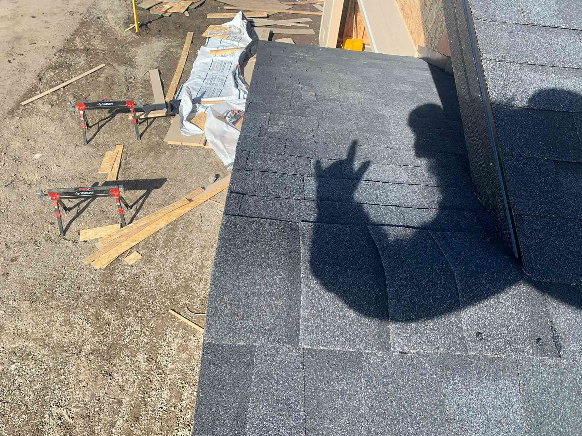 Roofing shingles on a flat roof beside tools, wood scraps, and a shadow of a person working