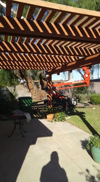 Backyard patio under wooden pergola, with a tree, chairs, potted plants, and a swing set in sunlight.