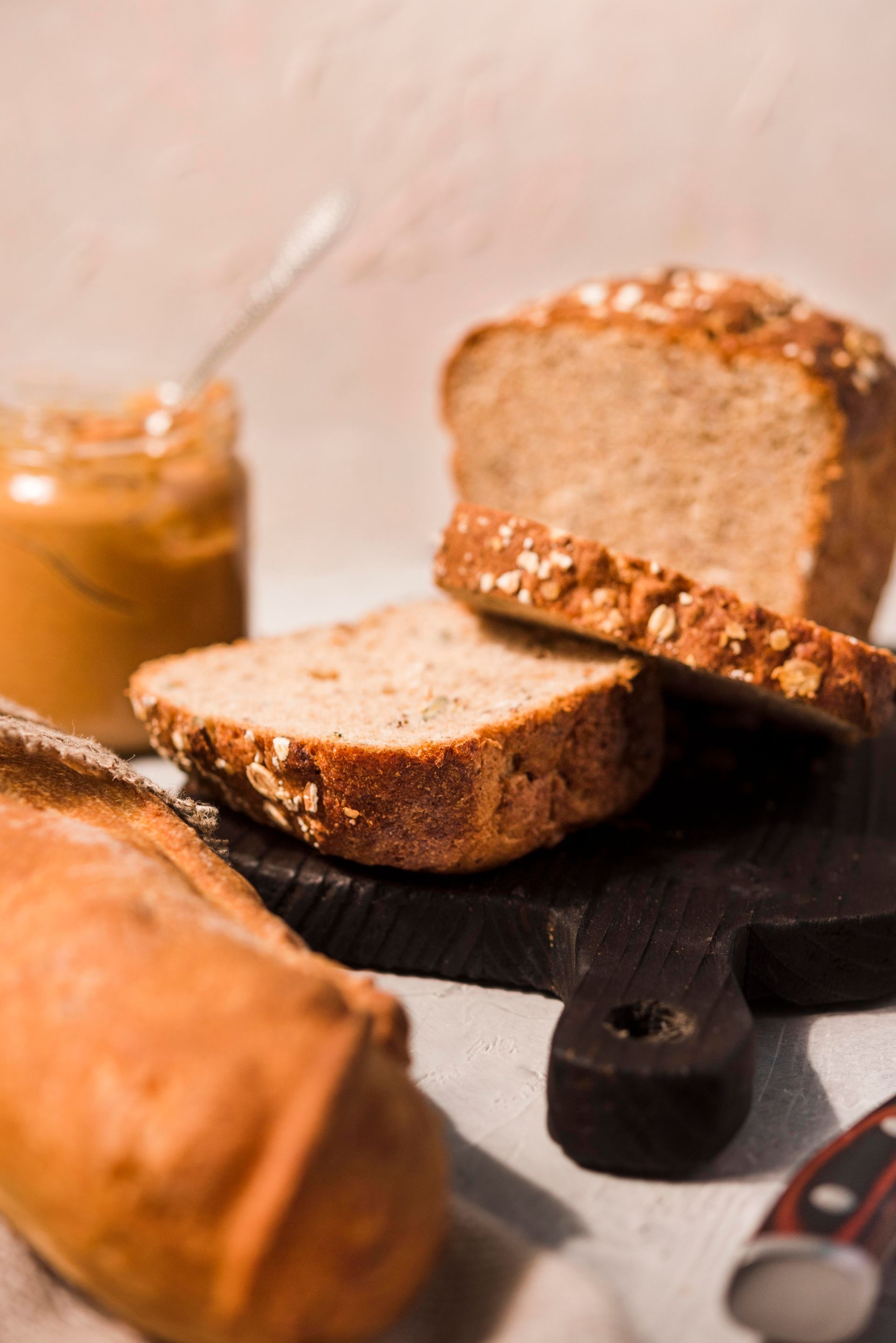 Pan de molde con avena, tarro de mantequilla de maní y una baguette sobre una tabla de madera.