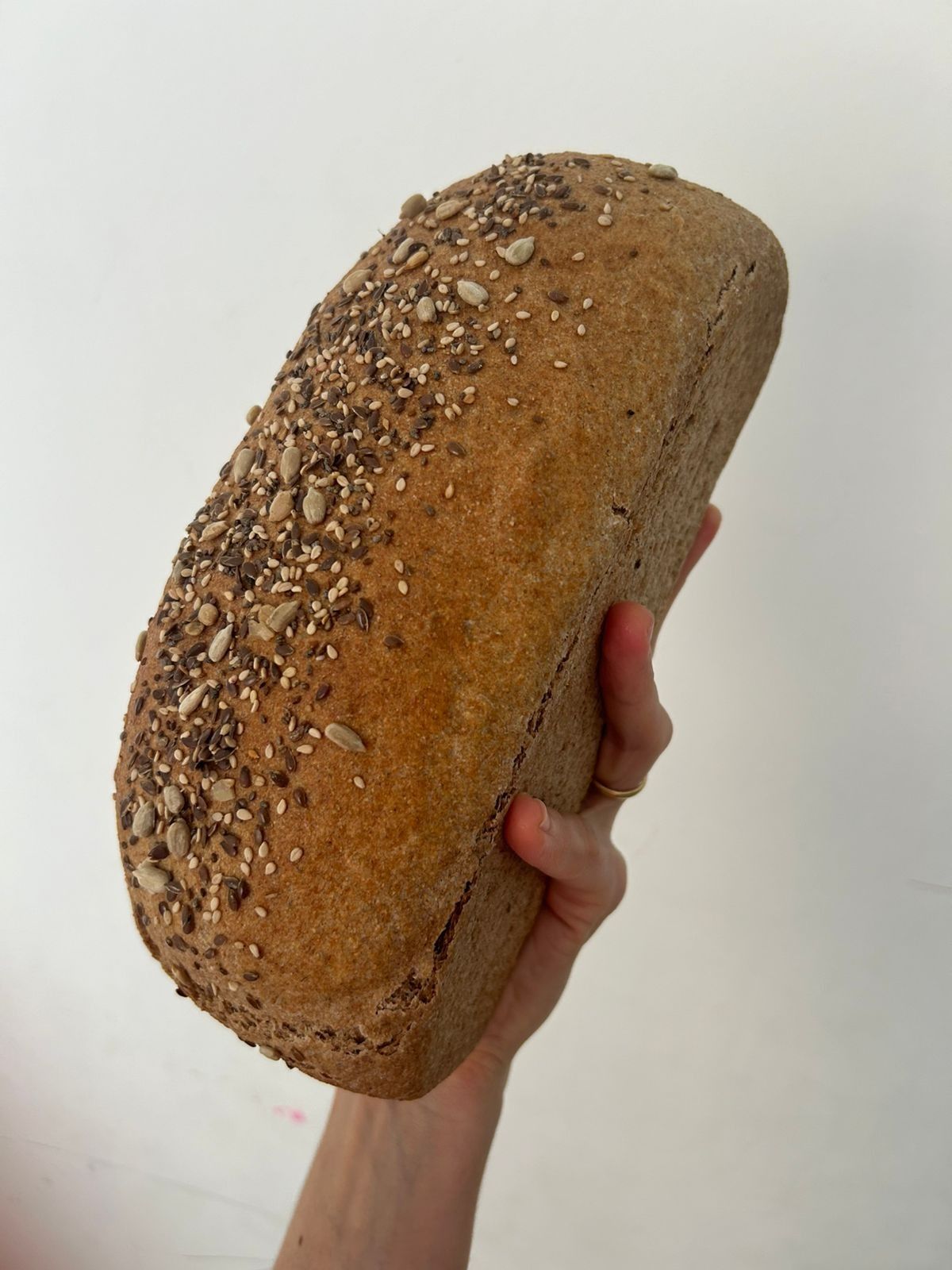 Mano sosteniendo una hogaza de pan integral cubierta de semillas sobre un fondo blanco.