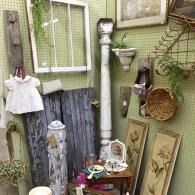 Small rustic outdoor display with framed art, hanging decor, and a white shirt on a chair against a green wall.