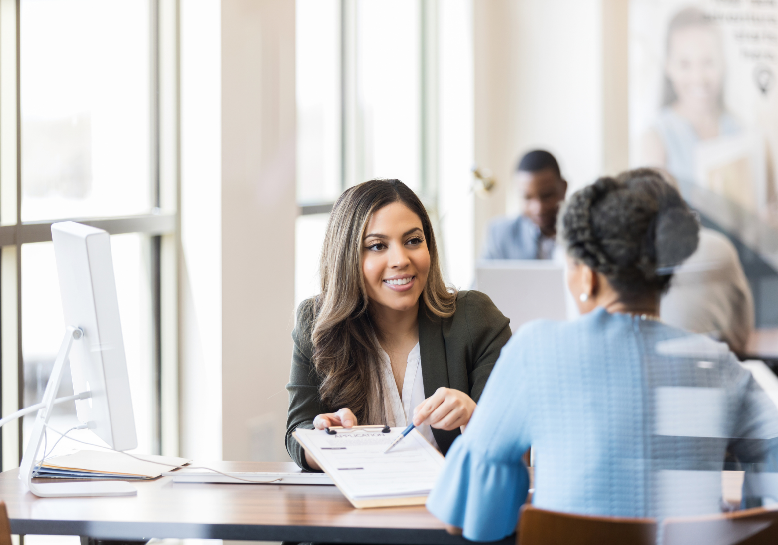 A professional in a blazer sits at a desk, pointing to a document for a colleague in a blue top in a bright office.