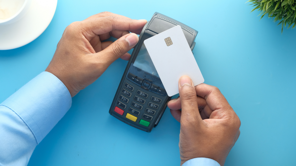 A person holding a blank white credit card over a payment terminal on a light blue surface.