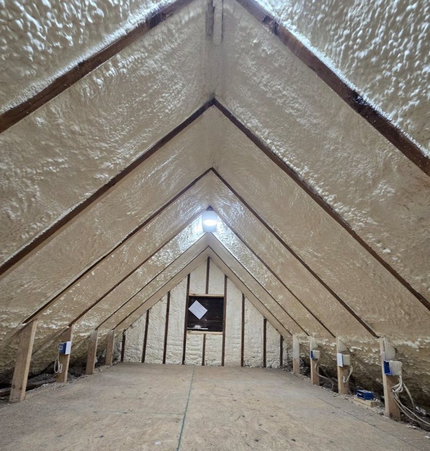 Attic interior fully insulated with light-colored spray foam, covering rafters and gable wall.