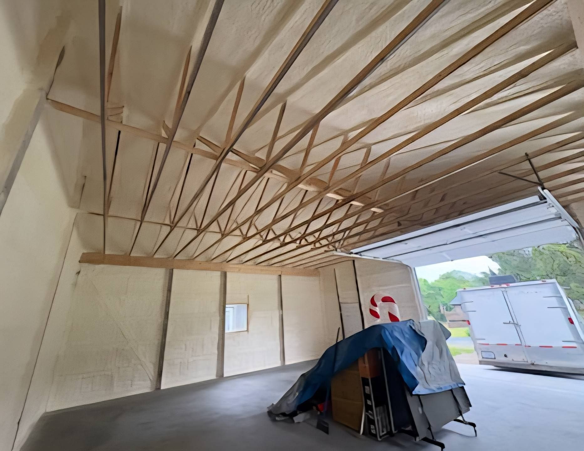 Interior of a garage with spray foam insulation on walls and ceiling; wood beams, and a partially open garage door.