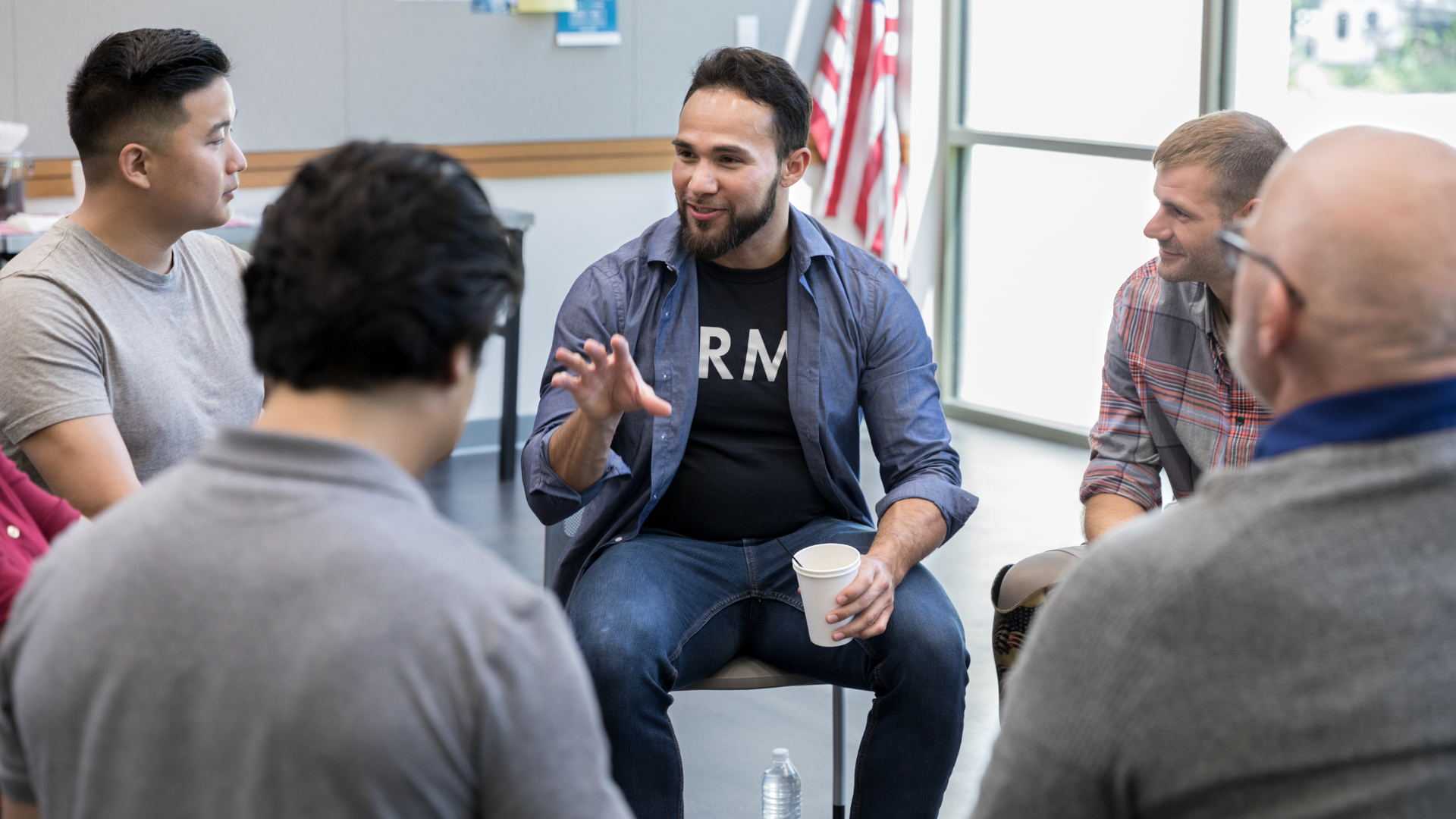 A group of men in a circle having a discussion, one gesturing with a cup in hand.