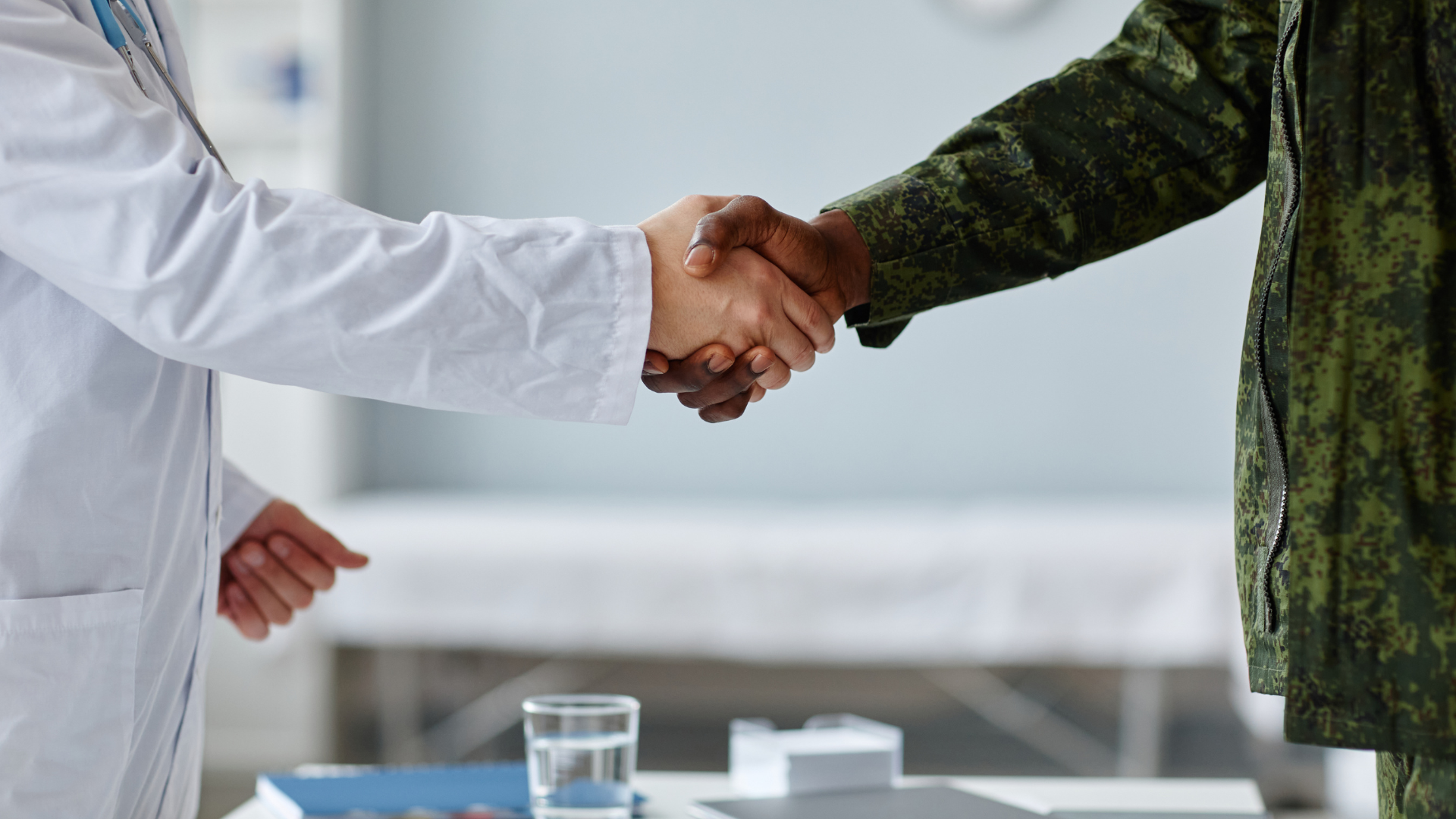 Doctor in white coat shakes hands with a person in military fatigues in a medical setting.