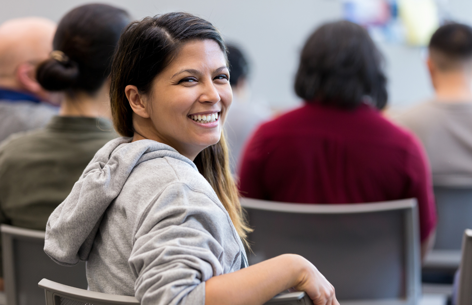 Woman in a gray hoodie smiles over her shoulder at the camera, seated in a room with other people.
