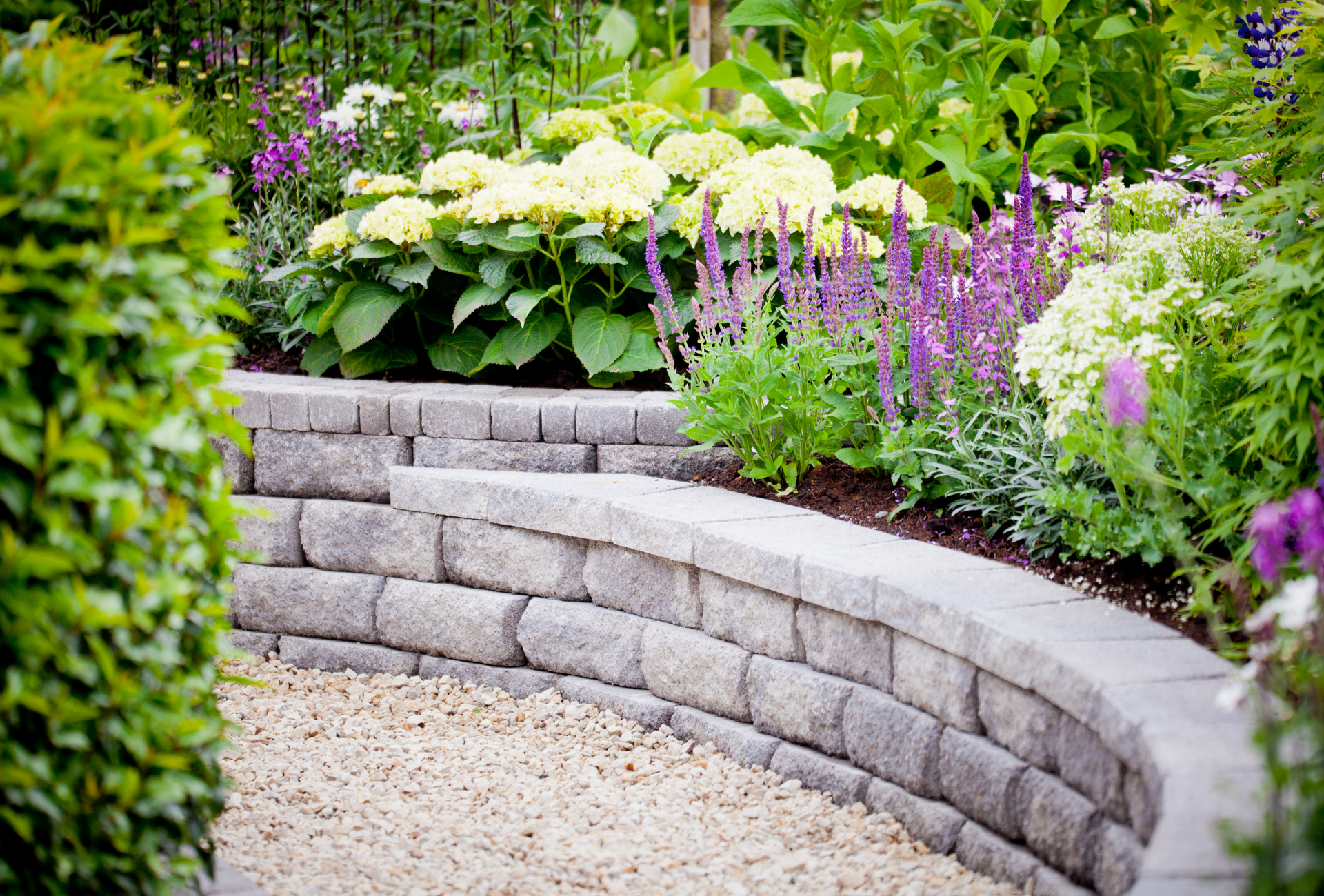 A stone wall in a garden surrounded by flowers and bushes.