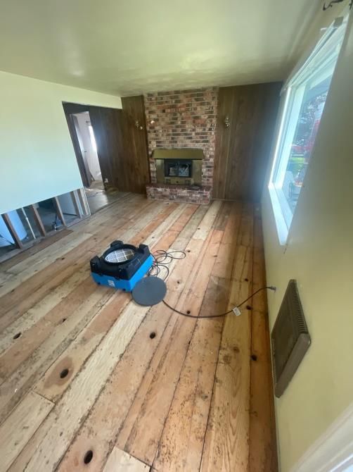 Room with exposed wood floor and brick fireplace, drying equipment in use, and partial wall removal.