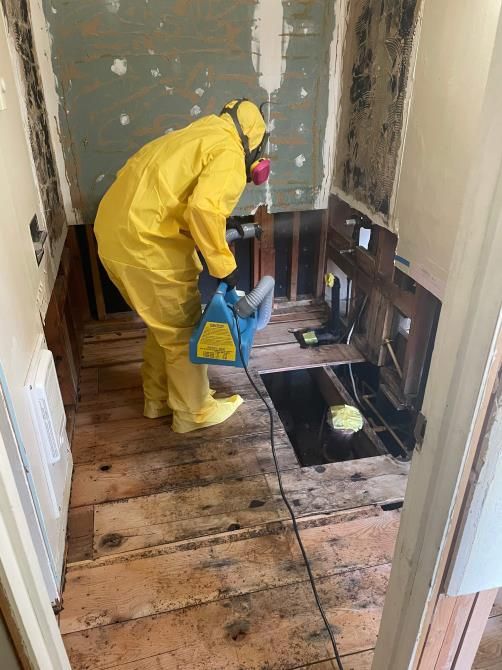 Person in hazmat suit removing mold from a bathroom; walls and floor are damaged.