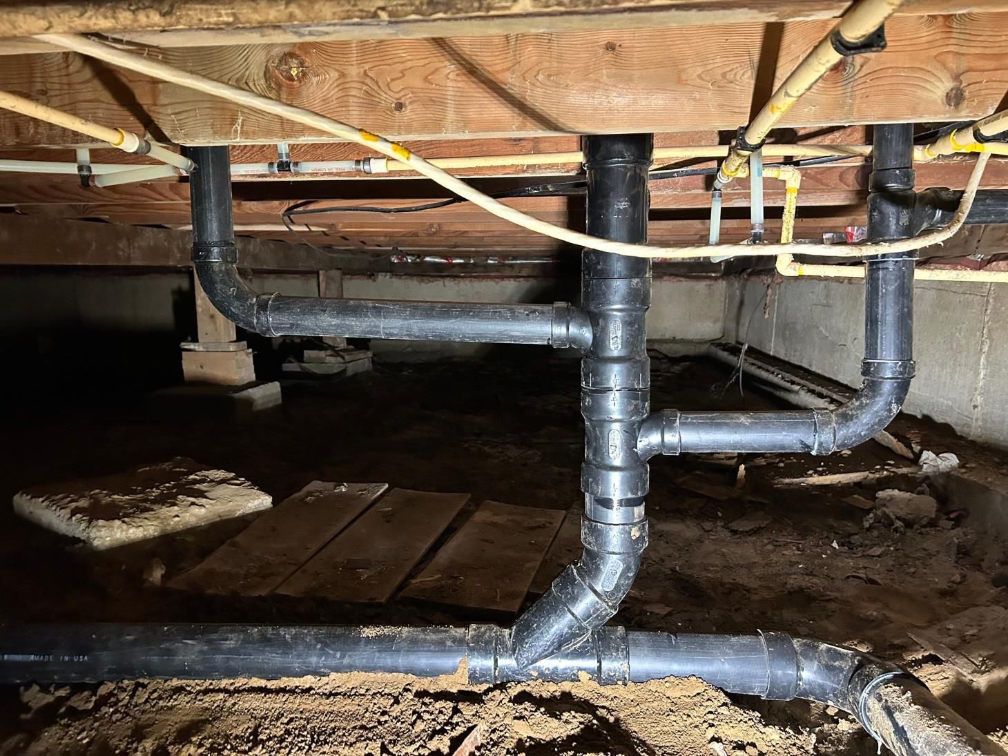 Black plumbing pipes installed under a wooden house, connecting to existing pipes.