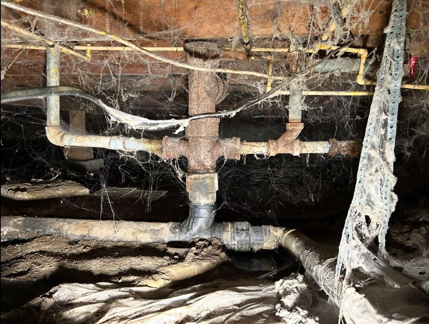 Rusty plumbing pipes covered in cobwebs in a dark crawlspace.